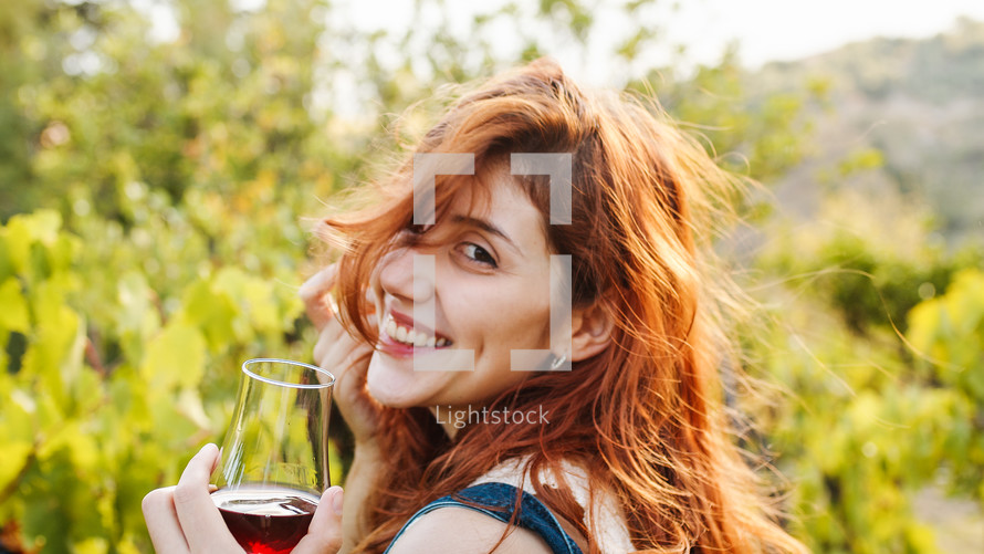 Beautiful red hair girl smile and walk with glass of wine among vineyards