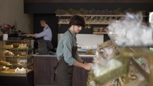 Father and son working in family bakery store. Son straightens goods on the shelf while father turns on coffee machine. Preparing for opening and visiting customers.