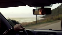 Bearded man driving a car along the coast, focused gaze in rearview mirror, golden hour.