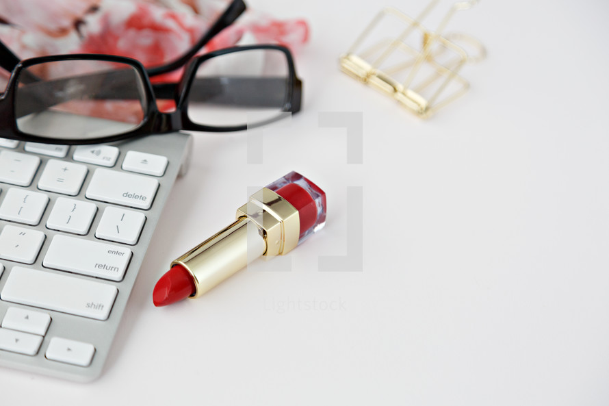 red lipstick, computer keyboard, floral scarf, gold clips, and reading glasses on a desk 
