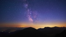 Timelapse of the Milky Way, the silhouette of the mountain landscape, the night city shining in the valley