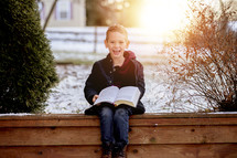 a child sitting outdoors in snow reading a Bible 