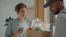 Young happy woman taking paper bag with groceries from delivery man, paying online with smartphone for order from supermarket, saying bye and closing door of apartment
