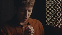 Young female parishioner with short haircut praying with hands clasped and eyes closed inside of confessional booth. Zoom-out shot
