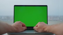 laptop with green screen male hands typing on keyboard man working at home office near the window