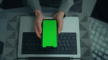 woman holding smartphone with green screen laptop on the desk
