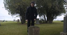 Young, sad man in black suit kneeling in cemetery at graveyard tombstone grieving in cinematic slow motion.
