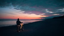 Girl with magic glowing bicycle walk near the ocean at sunset sky