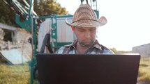 Farmer with laptop stood by tractor. Young farmer engineer with laptop standing on sun. This is a close up of a farmer using a computer on his tractor.