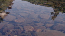 Close up dolly shot of the surface of a creek with smooth river rocks beneath the water reflecting the mountains.