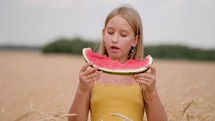 Girl eating juicy watermelon standing in wheat field on sunny summer day. 