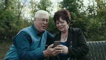 senior couple sitting on a park bench looking at a cellphone 