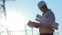 Foreman stands on construction site and holds drawings on paper in hands. Man shows hand where future construction be according to plan. Field with telecommunications tower. Male in protective helmet