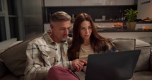 A serious middle-aged man with gray hair and a checkered shirt talks with his brunette wife while sharing a laptop in a modern kitchen on a sofa in a studio apartment