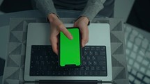 above view woman holding smartphone with green screen laptop on the desk
