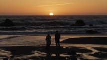 Couple watches sun set as waves crash on rocks, slow motion