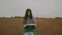 a woman walking through a field of tall grasses reading a Bible 