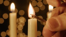 Person lighting a taper candle with a match with Christmas light bokeh in the background.