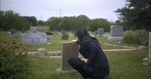 Young, sad man in black suit kneeling in cemetery at graveyard tombstone grieving in cinematic slow motion.