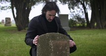 Young, sad man in black suit kneeling in cemetery at graveyard tombstone grieving in cinematic slow motion.