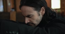 Young, emotional, anxious, and stressed man with long hair and black suit sitting in old church in worship and praying.