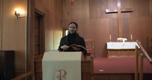 Young pastor at pulpit in black suit in old church building preaching, teaching sermon to congregation on Sunday morning worship service in cinematic slow motion in front of wood cross.