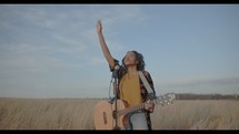 a woman with a guitar standing in a field with her arm raised 
