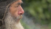 Side profile of a gray bearded man smoking a cigarette on the street.