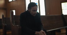 Young, emotional, anxious, and stressed man with long hair and black suit sitting in old church in worship and praying.