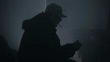 Silhouette of man wearing a hat and hooded coat in a dark, smoky room lighting and smoking a cigarette or drugs like marijuana. 