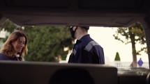 Beautiful woman and Delivery man in a protective mask and gloves opening the trunk and taking cardboard box parcel. Courier giving cardboard box to the male customer and closing minivan trunk with parcels. Footage from the backseat of the car.