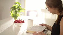 a woman sitting at a kitchen table reading a Bible 
