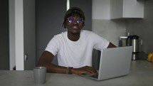 Young black male vlogger sitting with laptop at the kitchen, speaking on camera while livestreaming.