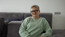 Senior woman smiling, looking to the camera, sitting on comfortable sofa.