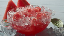 Close-up of a transparent glass bowl placed on ice cubes, filled with watermelon and ice. A woman's hand adds a sprig of mint into the dessert bowl