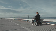 Wide shot of handicapped boy in electric wheelchair riding along the sea and enjoying fresh air
