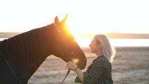 Blonde woman stroking and hugging horse. Beautiful lady with black stallion enjoying sunset nature. Love and friendship concept. Slow motion
