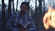 Man sitting beside a fire reading a book at dusk, enjoying nature
