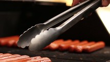 Hand with tongs moving hot dogs on a grill.