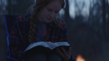 Young smiling woman sitting beside a camp fire and reading her Bible after sunset 