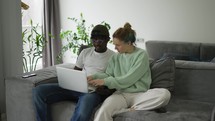 Biracial couple sitting on sofa with laptop, using smartphone for e-commerce.