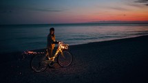 Girl with magic glowing bicycle walk near the ocean at sunset sky