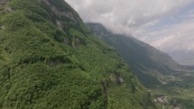 Cinematic drone advances, offering a view of dense greenery on a mountain slope with the tranquil valley visible below, all under a canopy of soft, drifting clouds