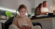 A little brown-haired girl with a braided hairstyle in a cream sweater sits and does something on her phone while her parents are preparing for a joint dinner in the evening