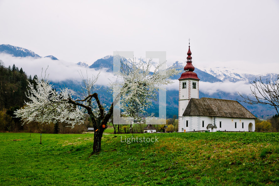 A white church with a red steeple in a green pasture and mountains.
