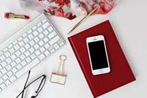 red lipstick, reading glasses, clip, computer keyboard, phone, scarf, pen, and red book on a desk 