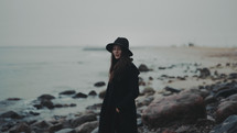 a woman in a trench coat and hat standing on a rocky shore 