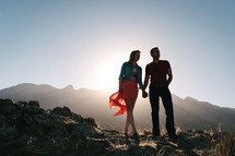 Silhouette of a man and woman, standing on a rocky hilltop, mountains in the background