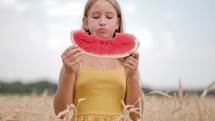 Cute girl eating watermelon standing in wheat field on sunny summer day. 