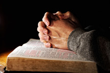 A man's hands clasped in deep prayer over a Holy Bible.